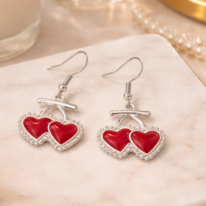 Red heart-shaped earrings on a marble surface with a candle and mirror in the background.