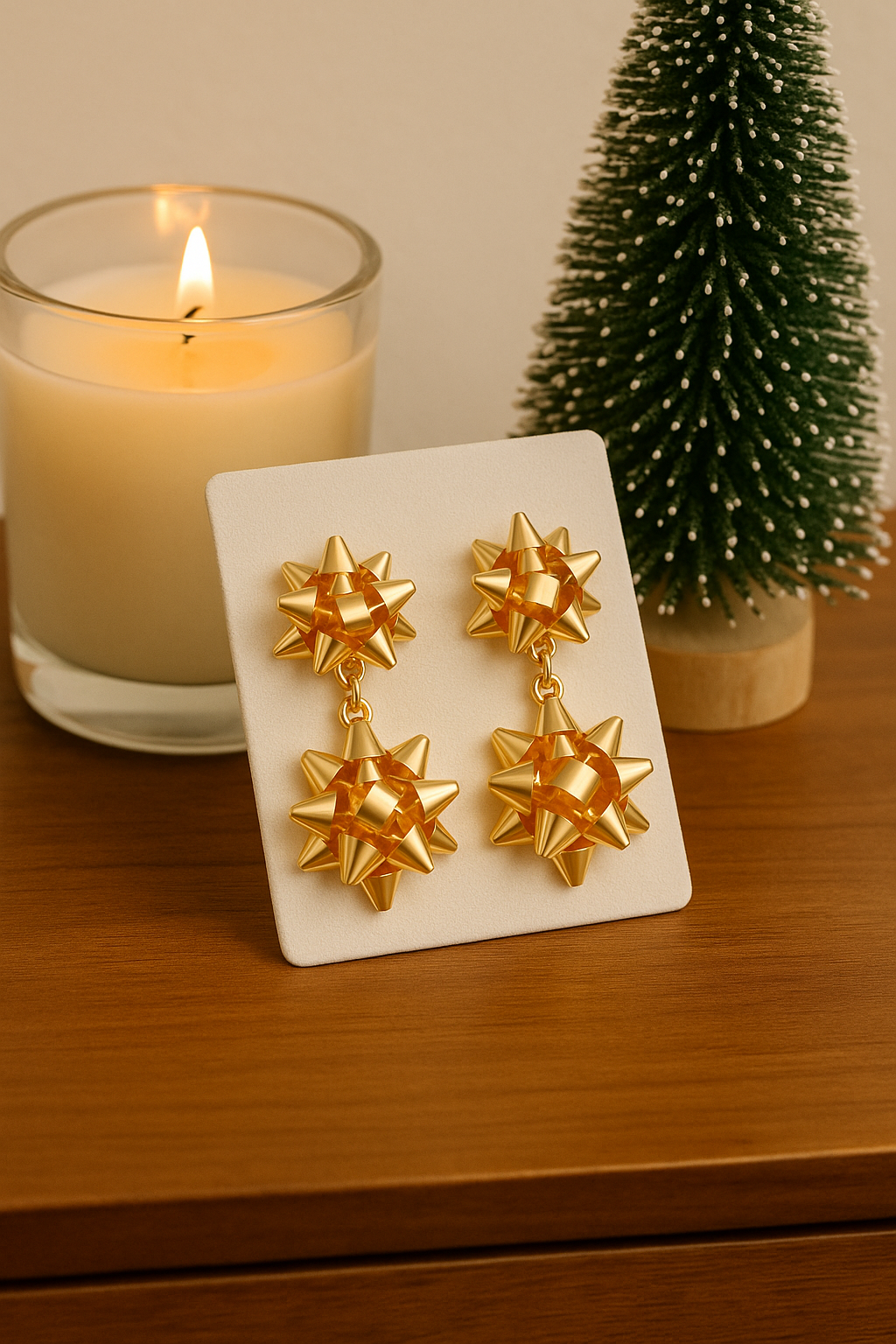 Gold bow-shaped earrings on a white card with a lit candle and small Christmas tree in the background.