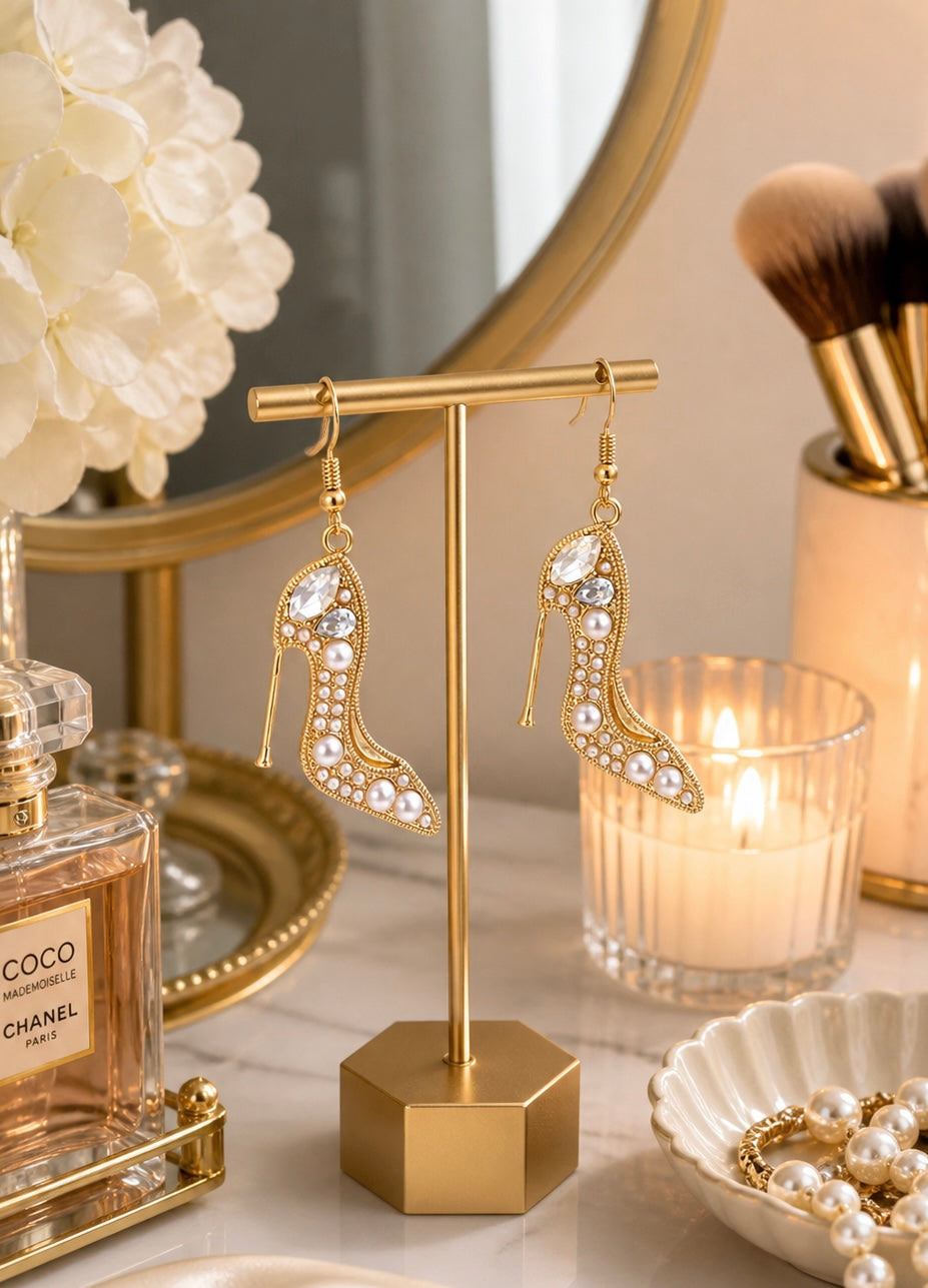 Gold high-heeled shoe earrings on a stand with a candle and perfume bottle in the background.
