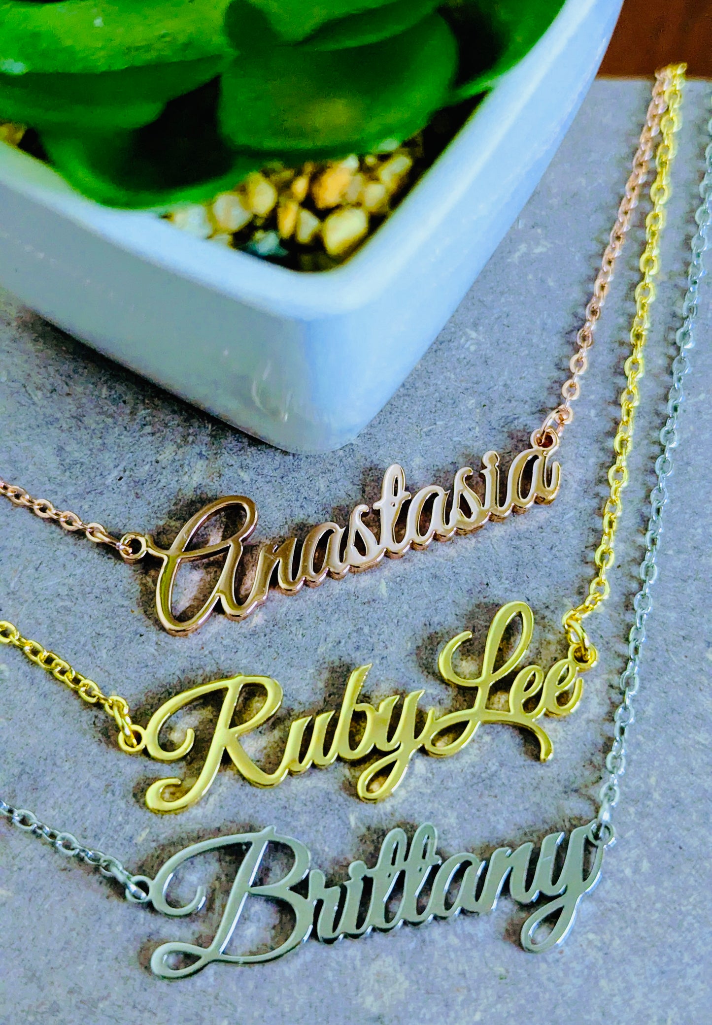 Gold nameplate necklaces on a stone surface with a bowl of greenery in the background.