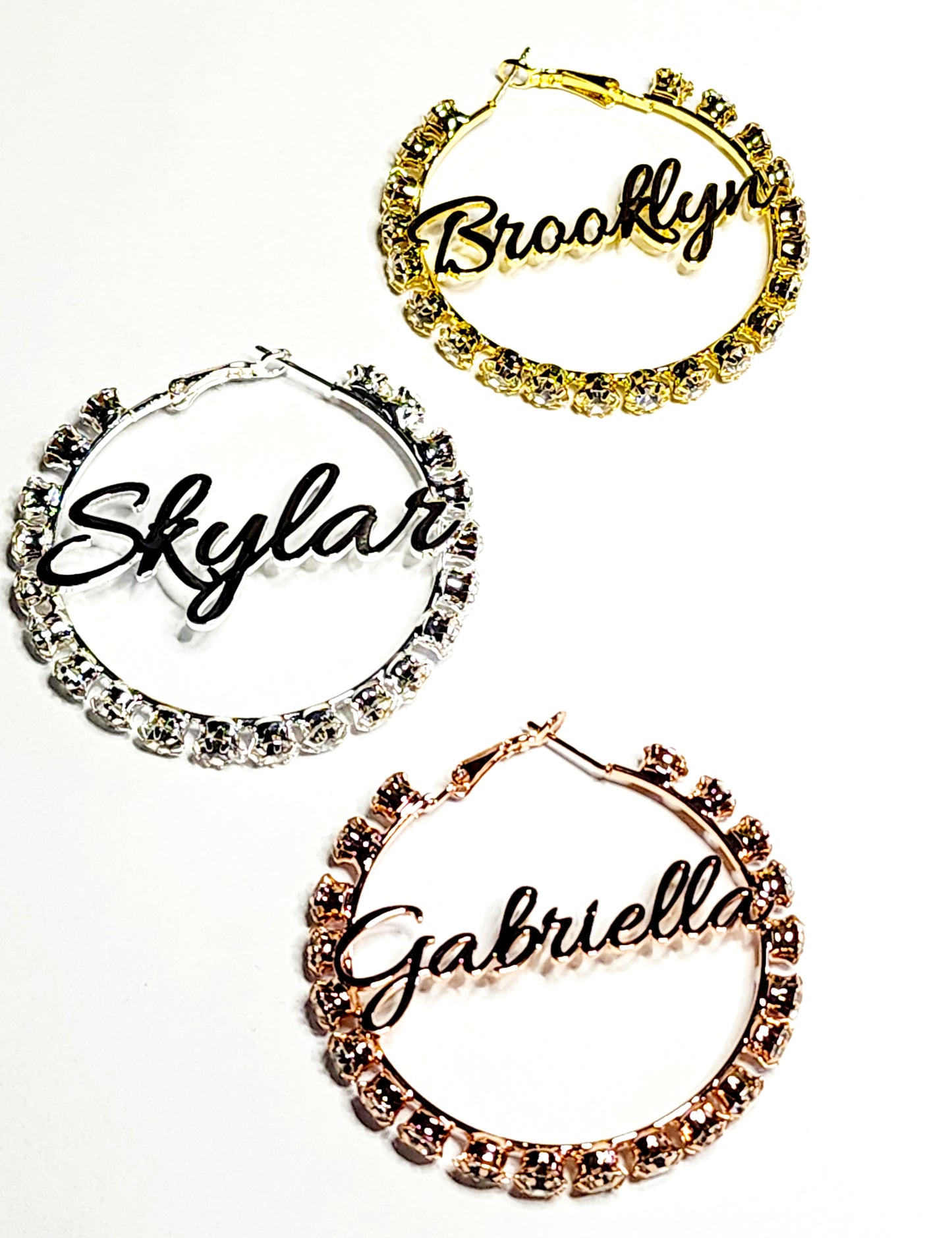 Three customized rhinestone hoop earrings in silver, gold, and rose gold colors, each with a different name written on them.