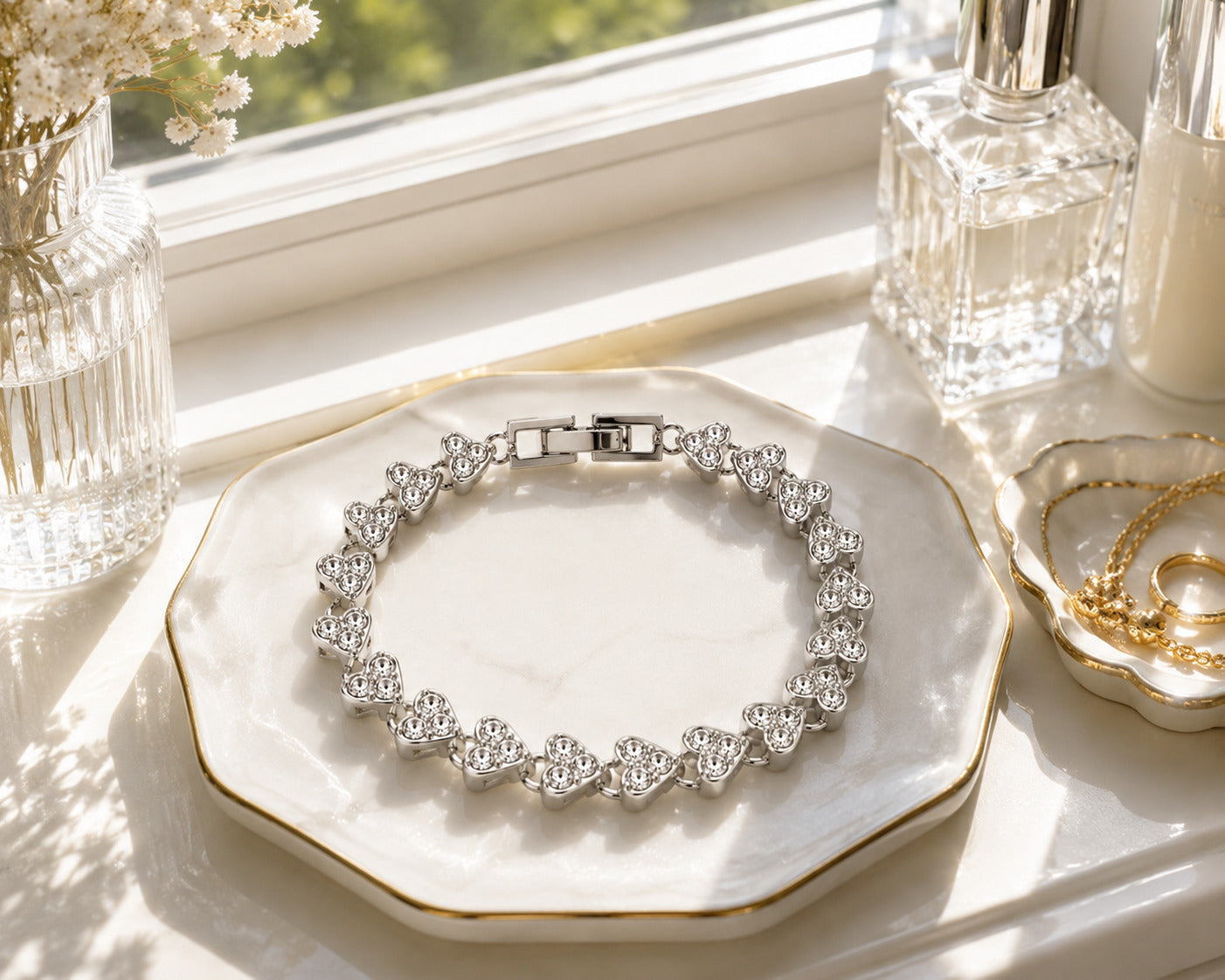 A silver-toned tennis bracelet with sparkling clear stones, displayed on a white ceramic plate with gold trim on a windowsill.