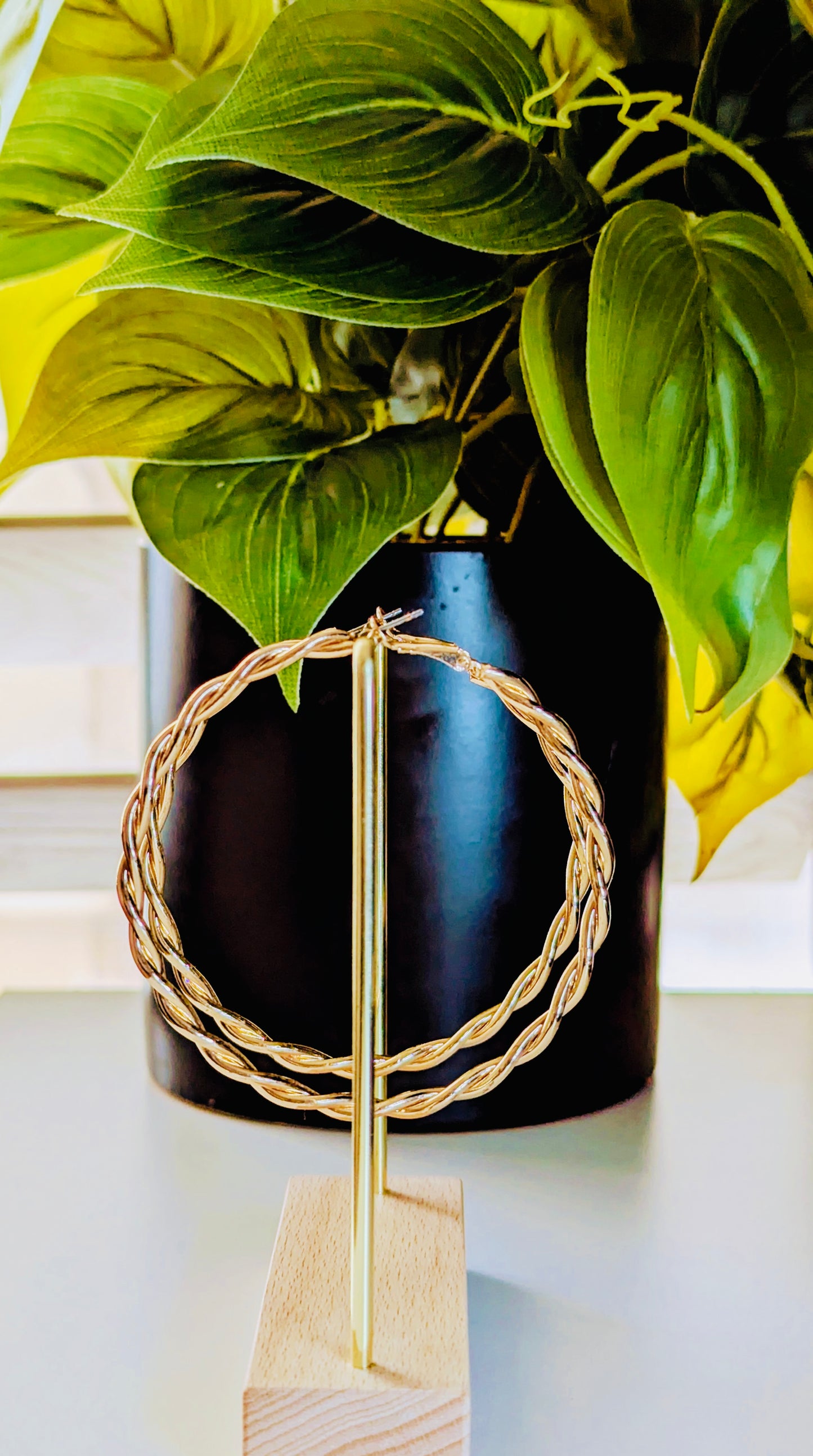 Decorative gold ring on a wooden stand with a black vase and green leaves in the background