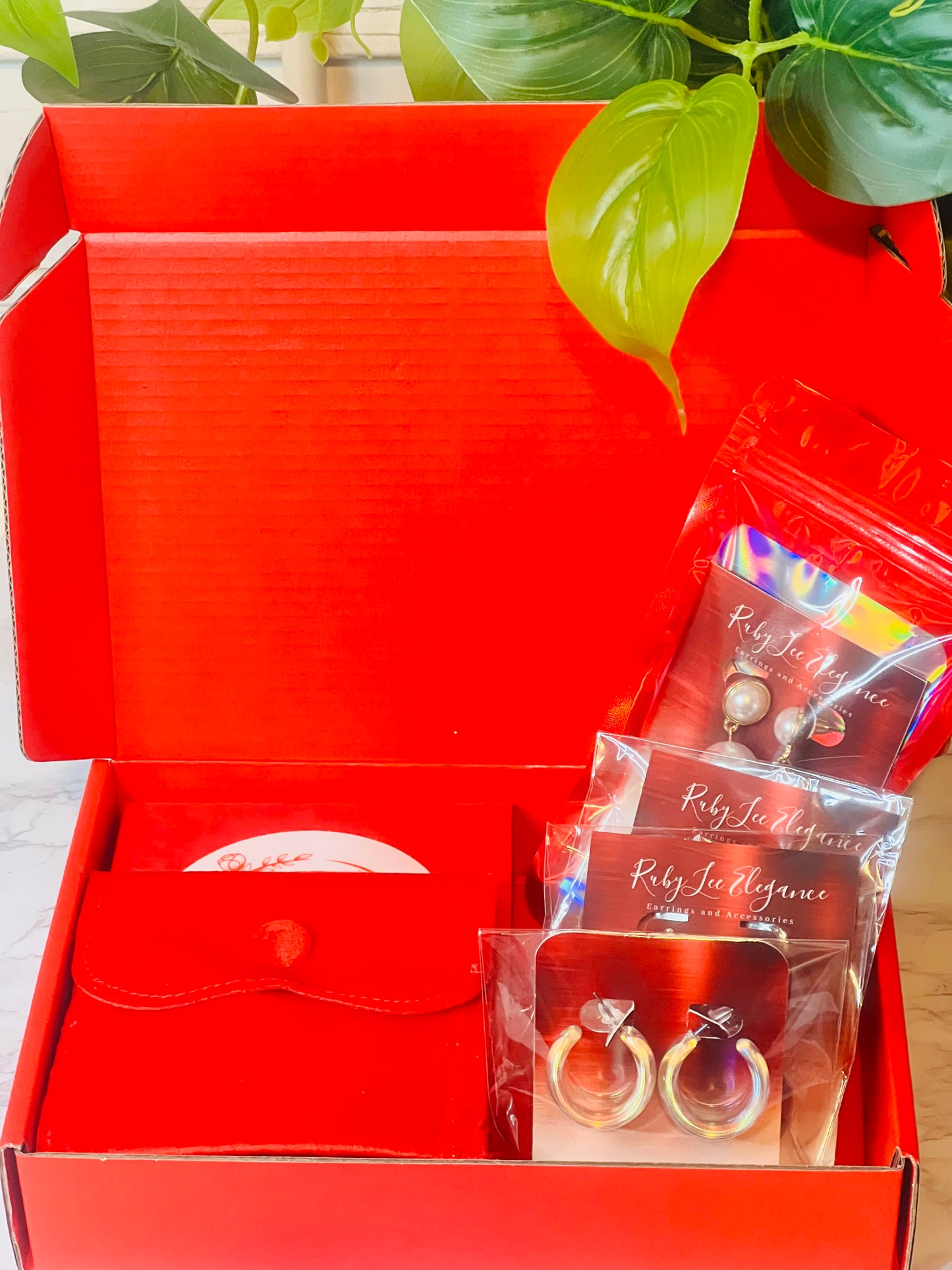 Red gift box with jewelry items including earrings and a card, with green leaves in the background.