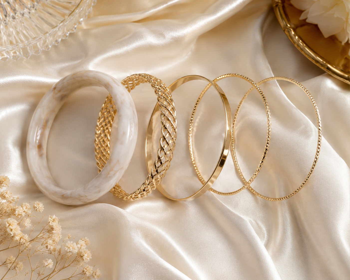 Five bangles arranged on draped satin fabric: one translucent white bangle with subtle marbling, one gold bangle with a braided texture, and three gold bangles with smooth, polished finishes.