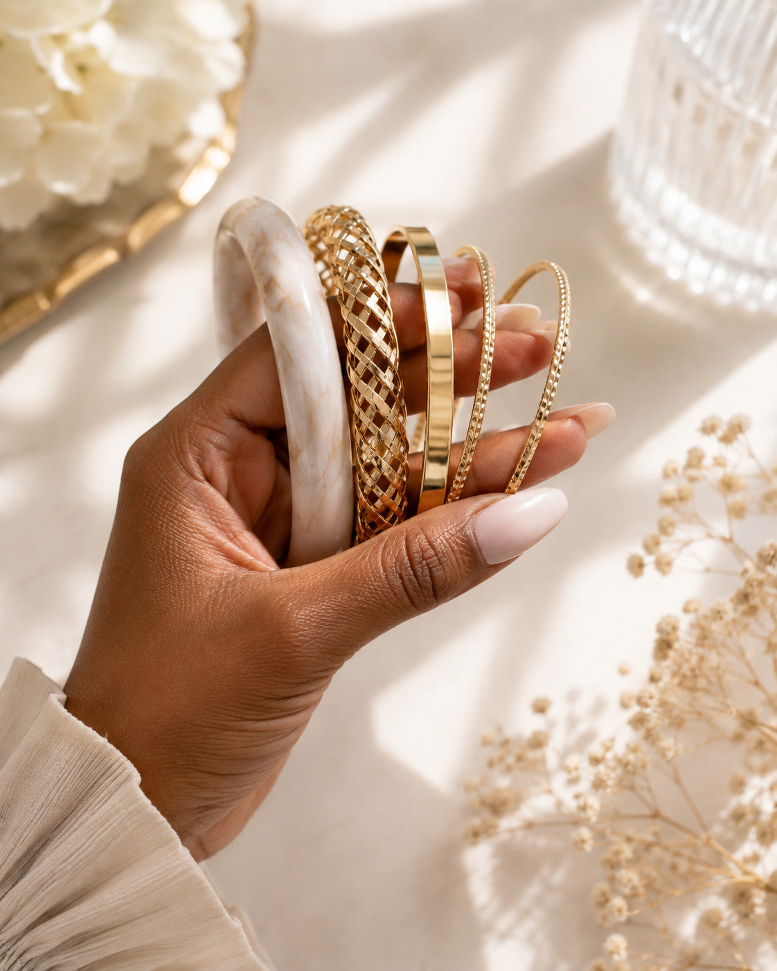 A hand holding a stack of five bangles, including a white marble bangle, a gold lattice bangle, a gold bar bangle, and two gold chain bangles, styled on a warm beige surface with dried flowers.