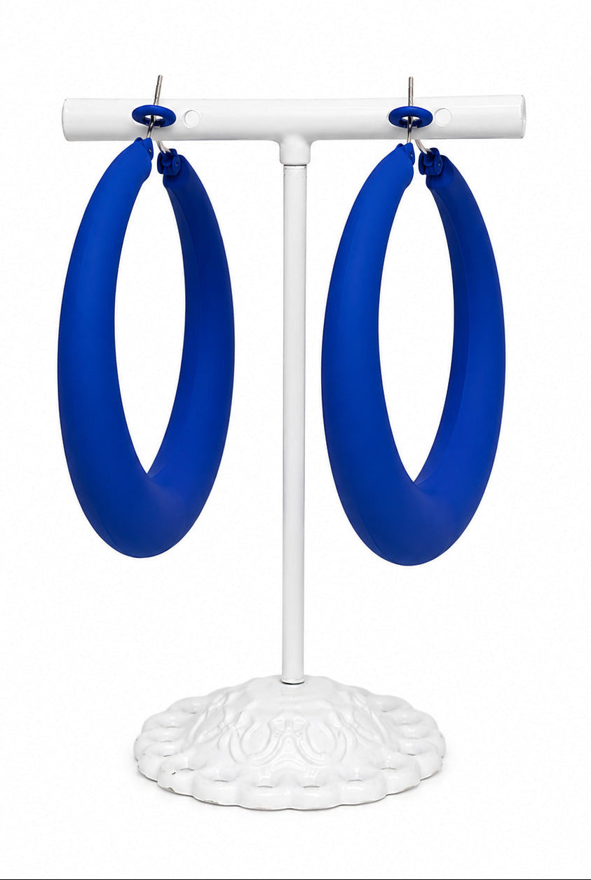 A pair of large, vibrant blue hoop earrings displayed on a white metal stand against a solid white background.