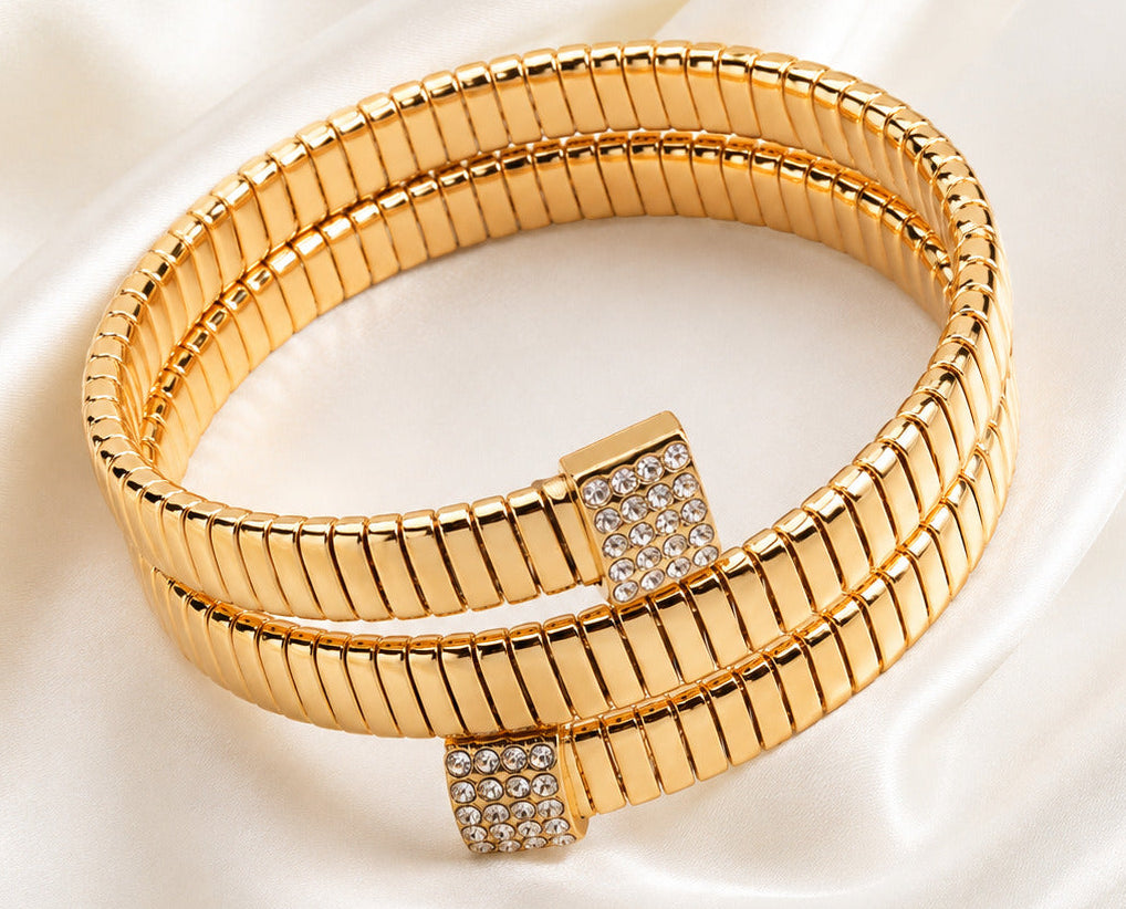 A double-strand gold bangle bracelet with a textured, ribbed design and square crystal-encrusted clasps, displayed on a draped white satin fabric.