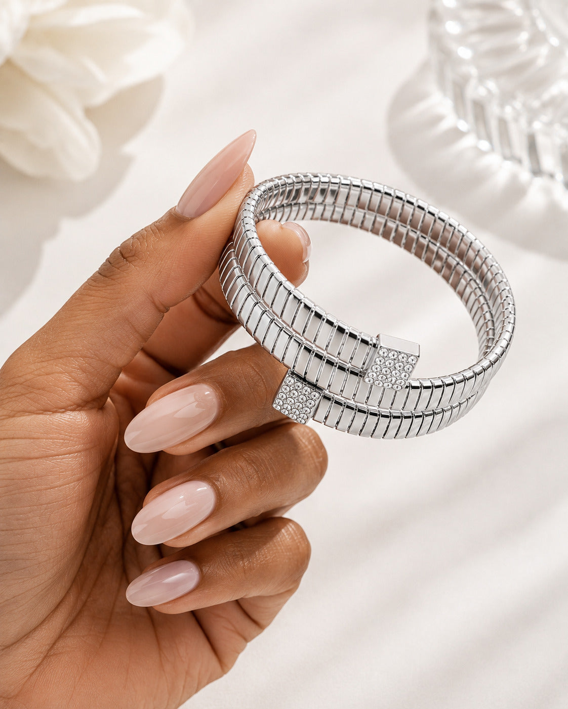 A close-up of a hand holding a silver-toned multi-band bracelet with a textured, snake-like segmented design and pavé-set accents on the clasp, set against a soft white background with blurred decorative elements.