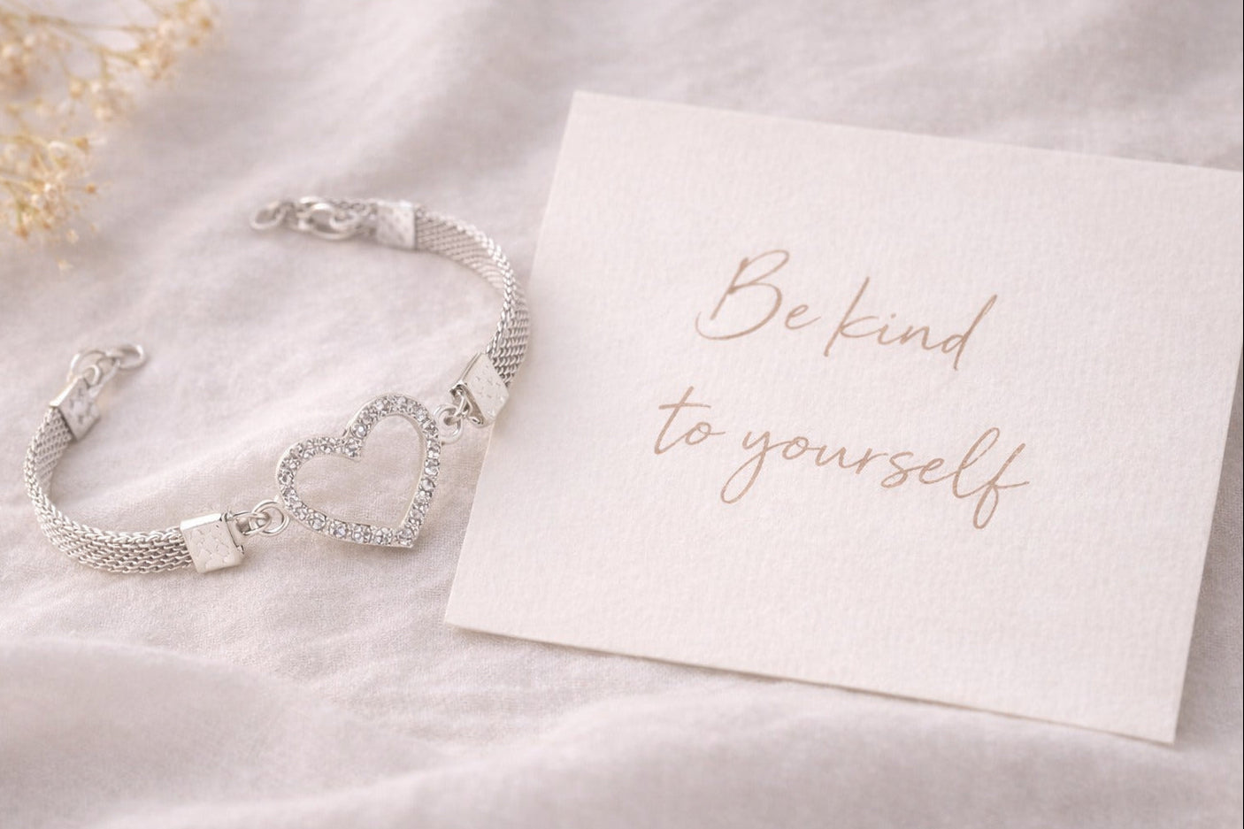 Silver bracelet with heart design on a white card with 'Be kind to yourself' text, on a soft white background.