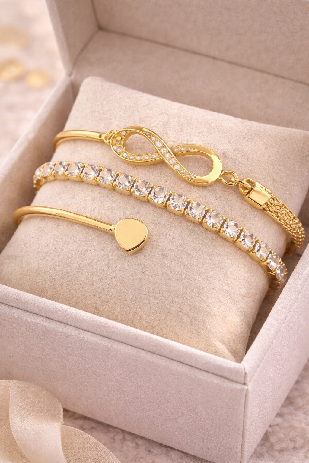 Gold bracelets with heart and infinity designs on a beige cushion in a jewelry box.