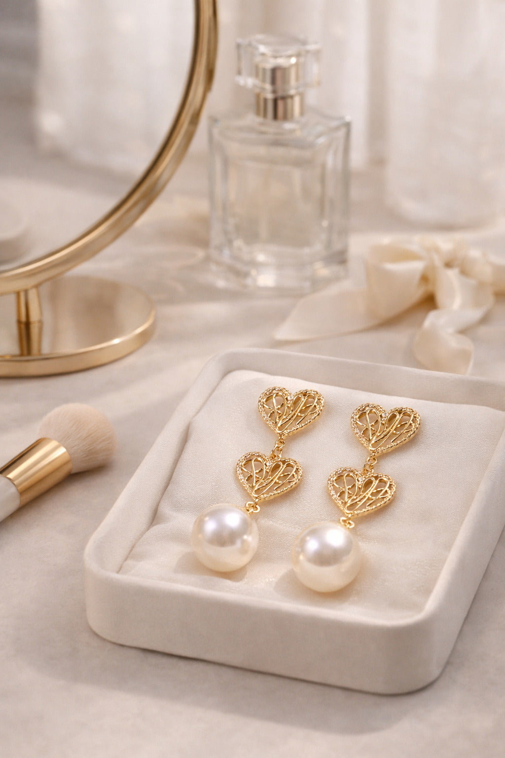 Gold heart-shaped earrings with pearls on a white jewelry box, next to a mirror and perfume bottle.