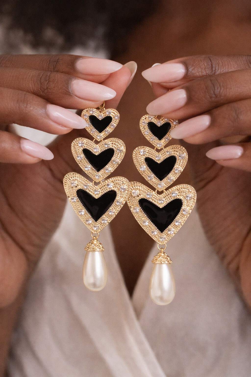 Heart-shaped earrings with black centers and pearl drops held by a person with light pink nail polish.