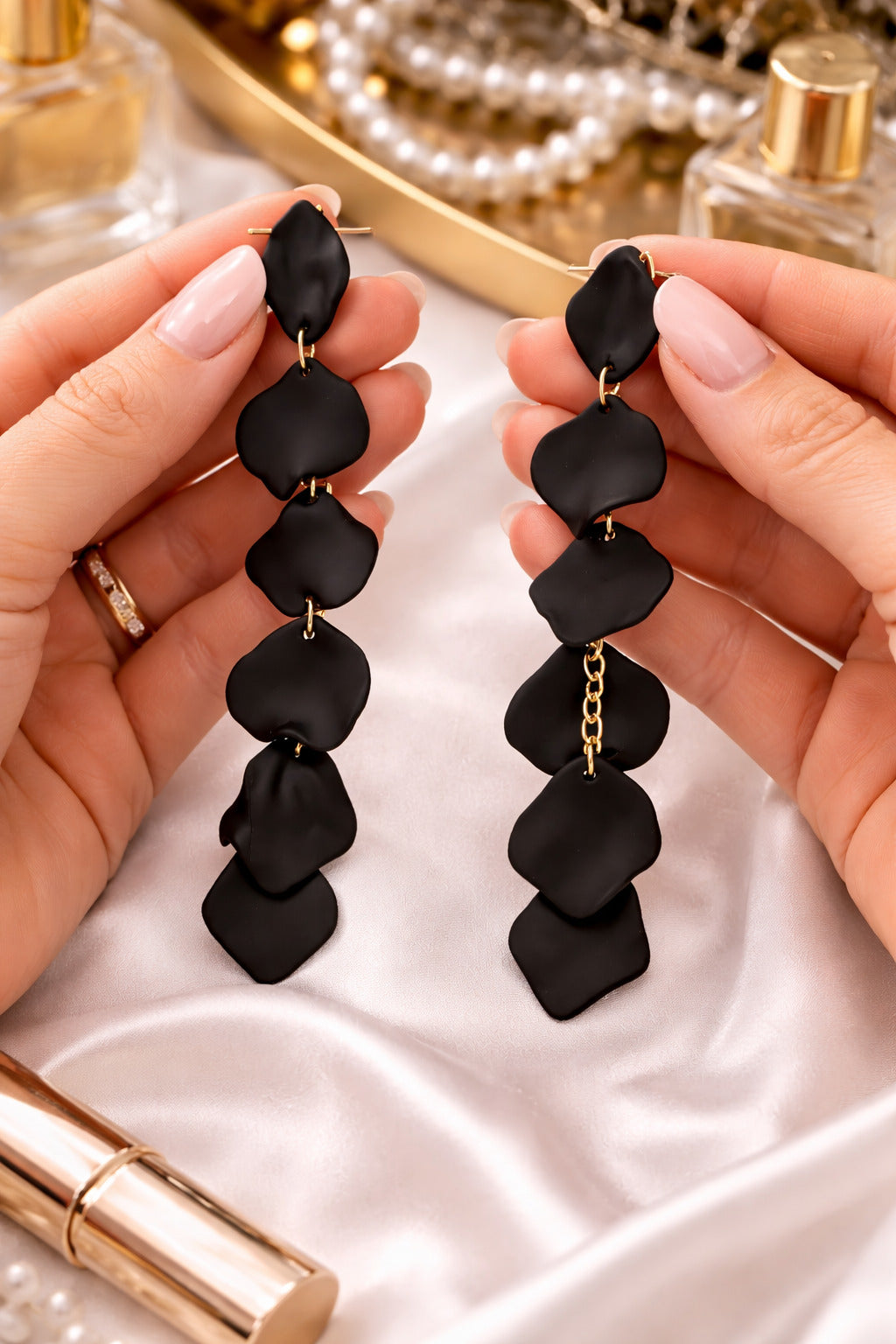 Black statement earrings held by hands with a blurred background of perfume bottles and jewelry.