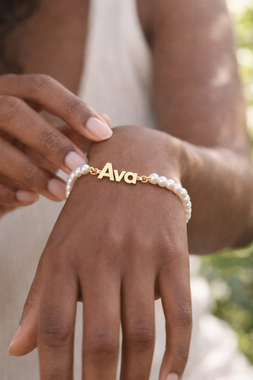 Hand wearing a gold 'Ava' bracelet with a pearl bracelet against a blurred natural background