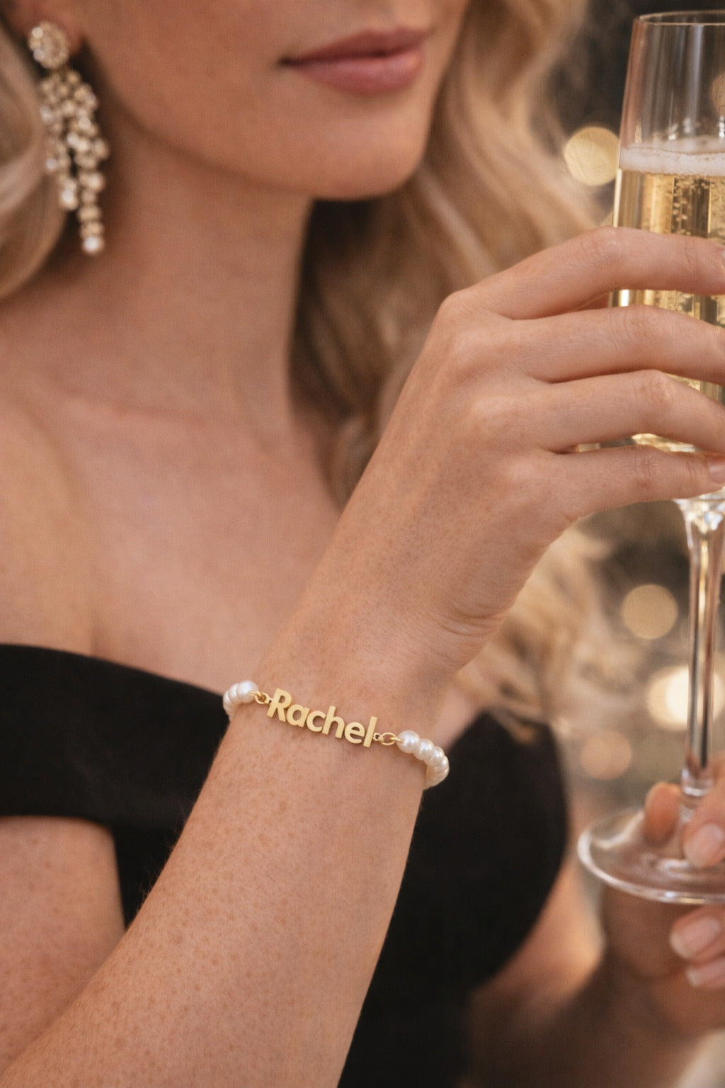 Woman holding a glass of champagne with a 'Rachel' bracelet on a blurred background