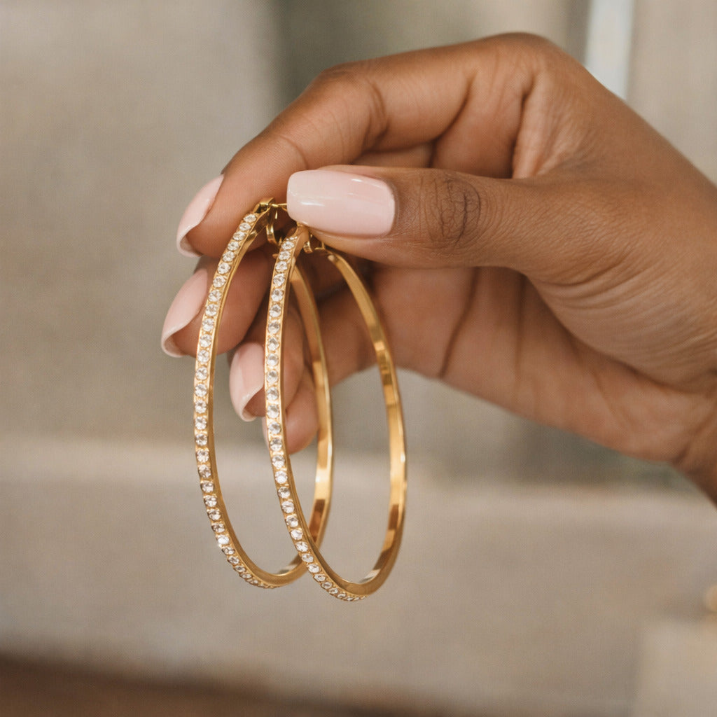 Gold hoop earrings held by a hand with light pink nail polish against a neutral background