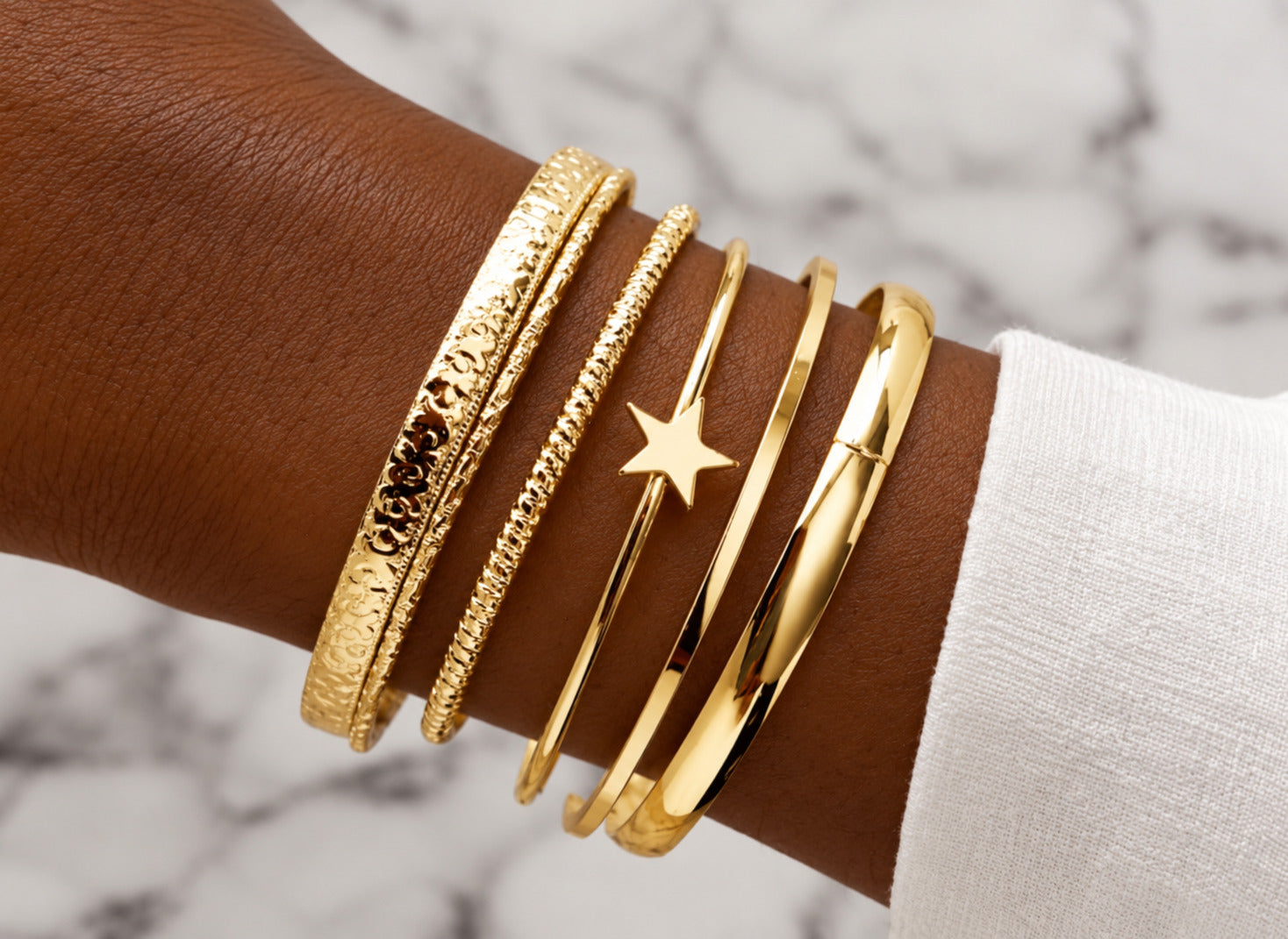 A close-up of a person's wrist wearing five stacked gold bangles, including textured, rope, and smooth bands with a star charm, set against a white marble background.