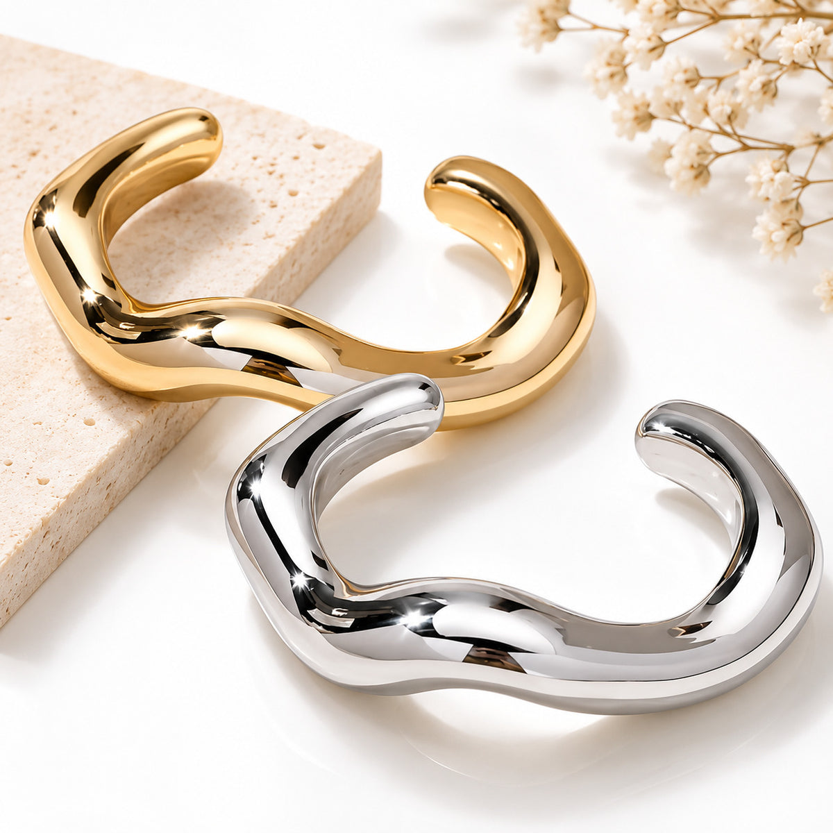 Two abstract wavy cuff bracelets, one polished gold and one polished silver, displayed on a white surface with a beige stone block and dried flowers.