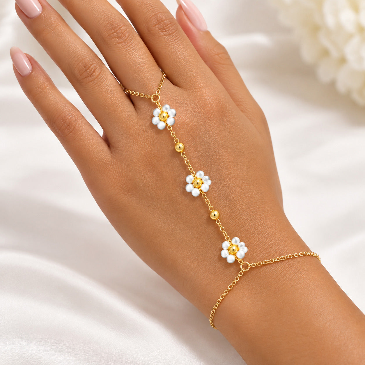 A gold chain hand bracelet with three white flower charms and pearl-like centers, shown worn on a person's hand against a soft white fabric background.
