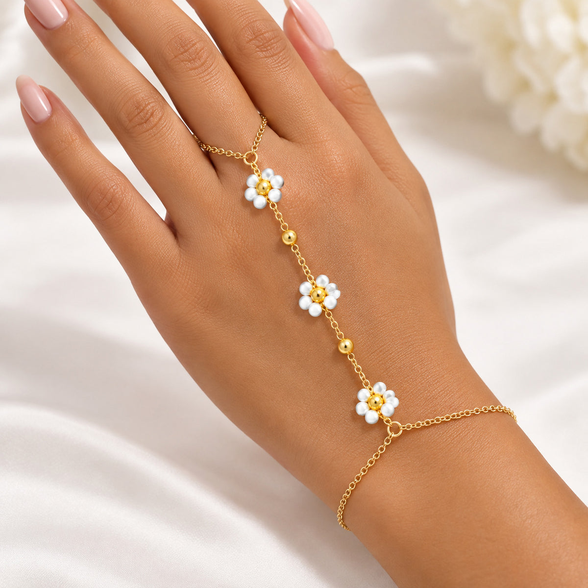 A gold chain hand bracelet with three white flower charms and pearl-like centers, shown worn on a person's hand against a soft white fabric background.