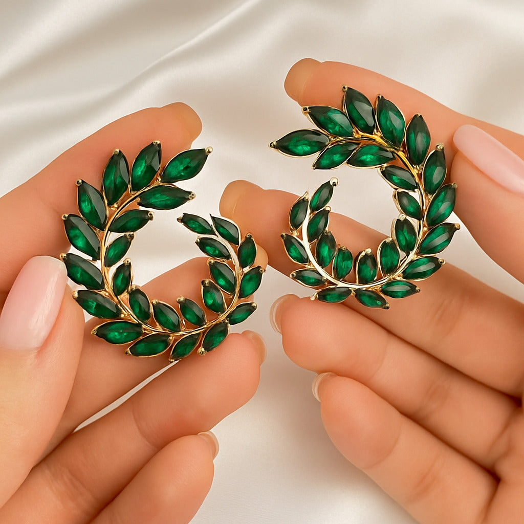Gold and green leaf-shaped earrings held between two hands against a neutral background