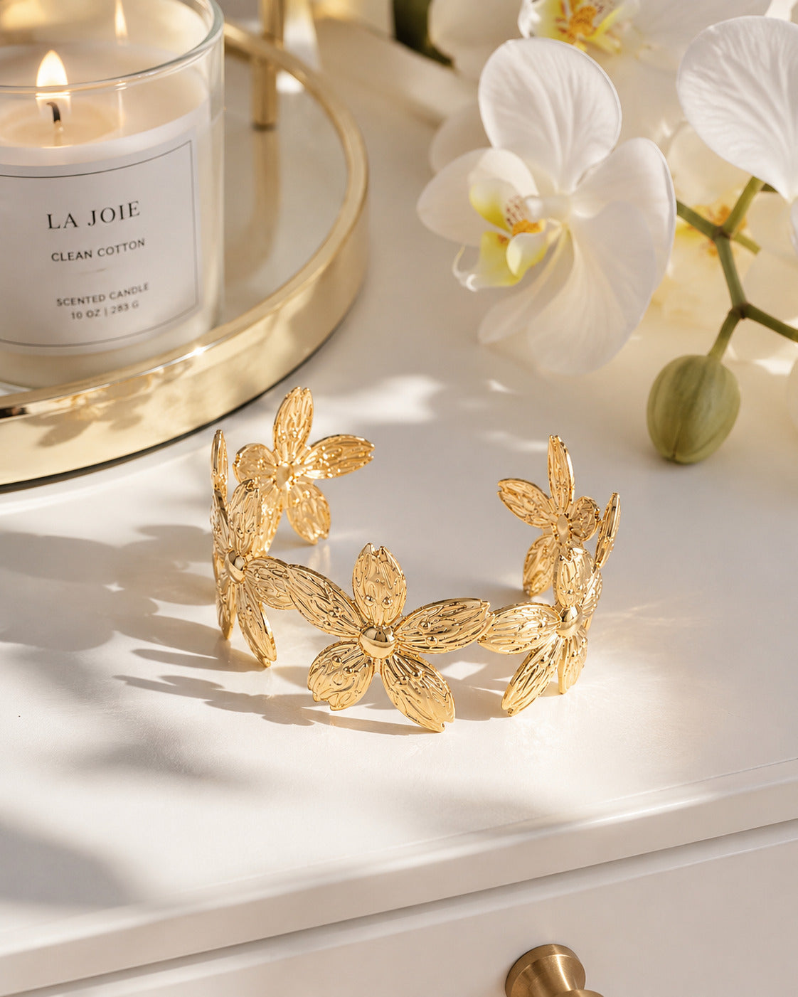 A gold cuff bracelet decorated with textured floral motifs, displayed on a white surface with warm sunlight and shadows. A lit candle in a glass jar on a gold tray and white orchids are in the background.