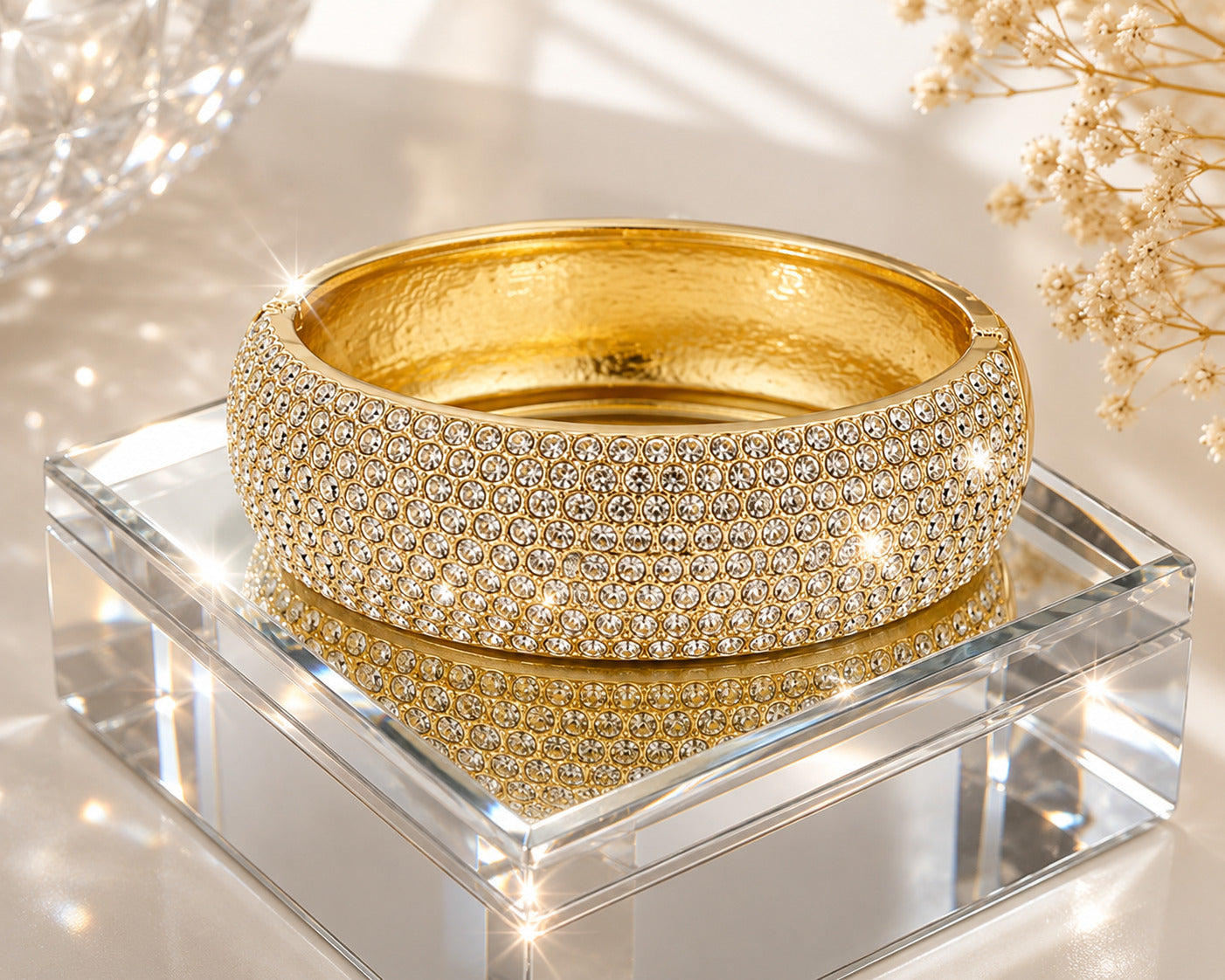 A wide gold bangle bracelet covered in rows of clear pavé stones, displayed on a clear acrylic block with a reflective surface.