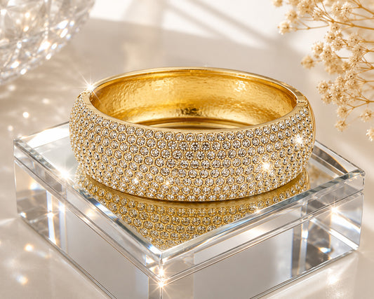 A wide gold bangle bracelet covered in rows of clear pavé stones, displayed on a clear acrylic block with a reflective surface.