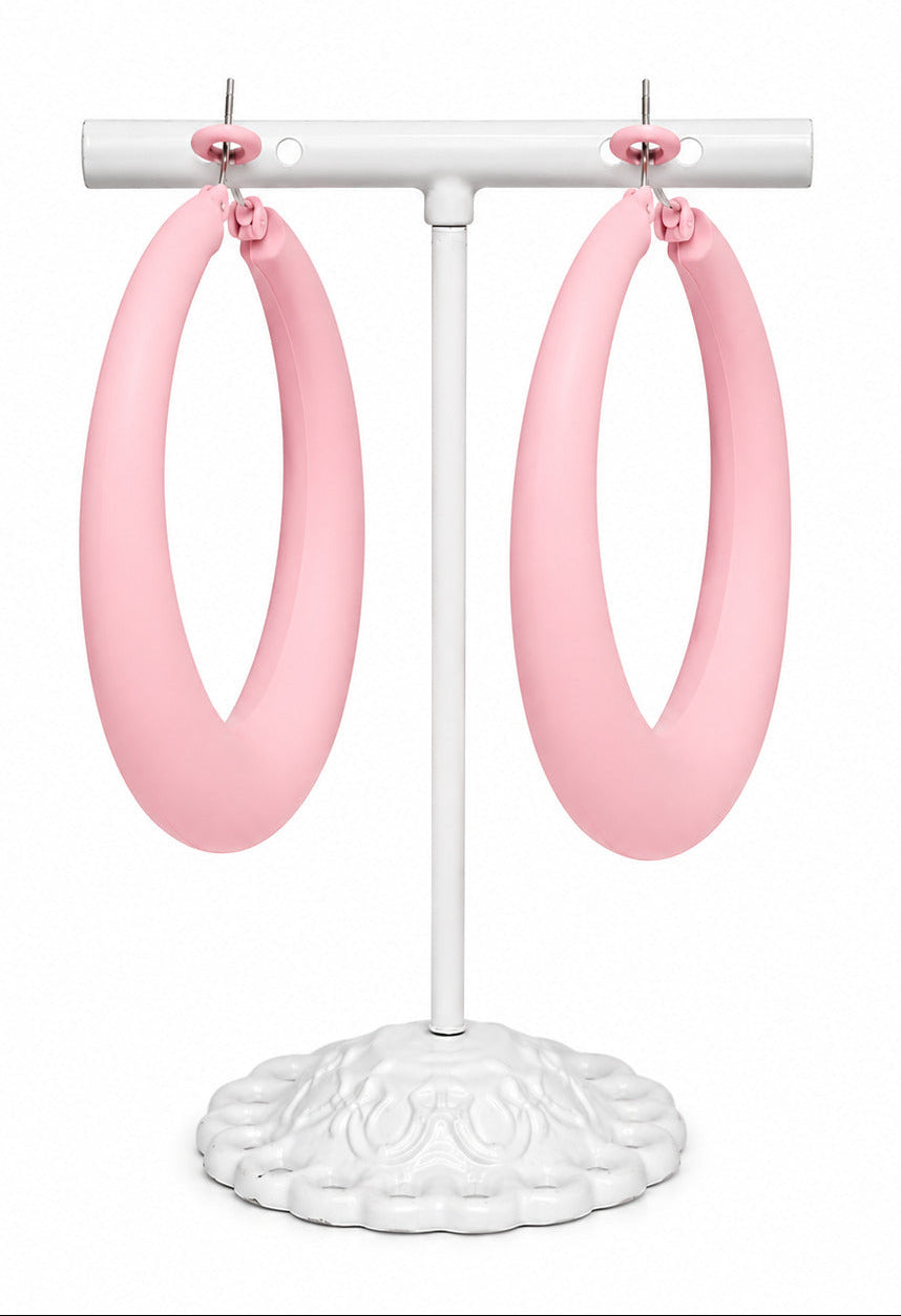 A pair of oversized light pink hoop earrings displayed on a white jewelry stand against a plain white background.