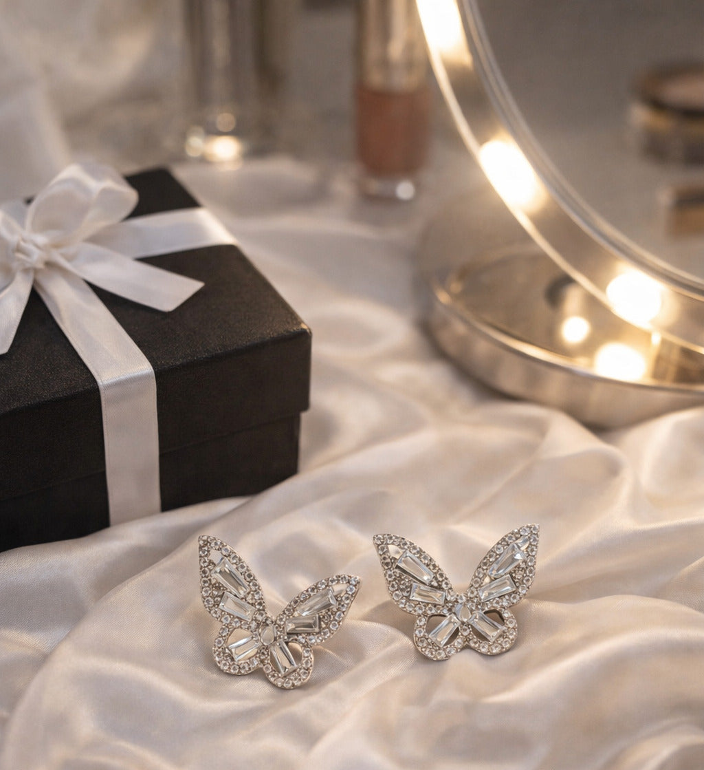 Butterfly-shaped earrings on a white fabric surface with a gift box and mirror in the background.