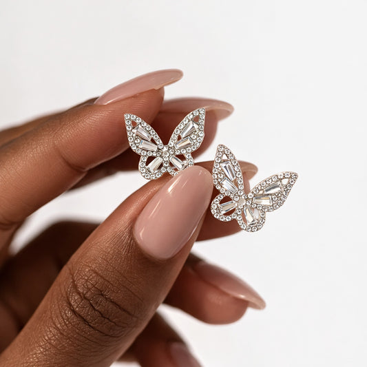 Butterfly-shaped earrings held by a hand with pink nail polish on a white background