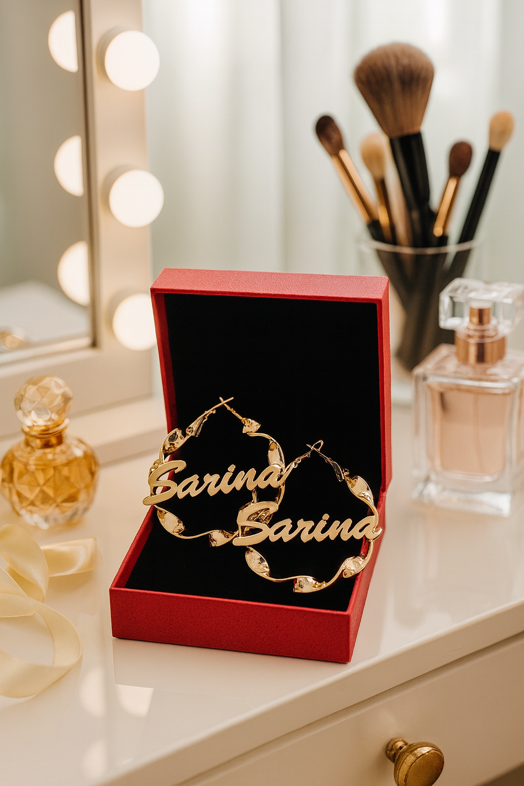 Gold 'Carina' earrings in a red box on a vanity with makeup brushes and perfume.