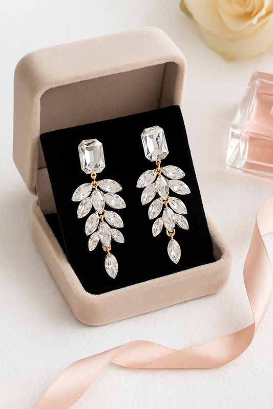 Crystal leaf earrings in a jewelry box with a pink ribbon and perfume bottle in the background.