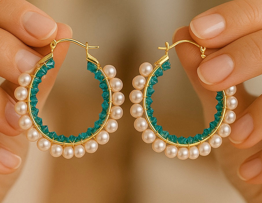 Gold hoop earrings with pearl and teal bead accents held by hands against a blurred background