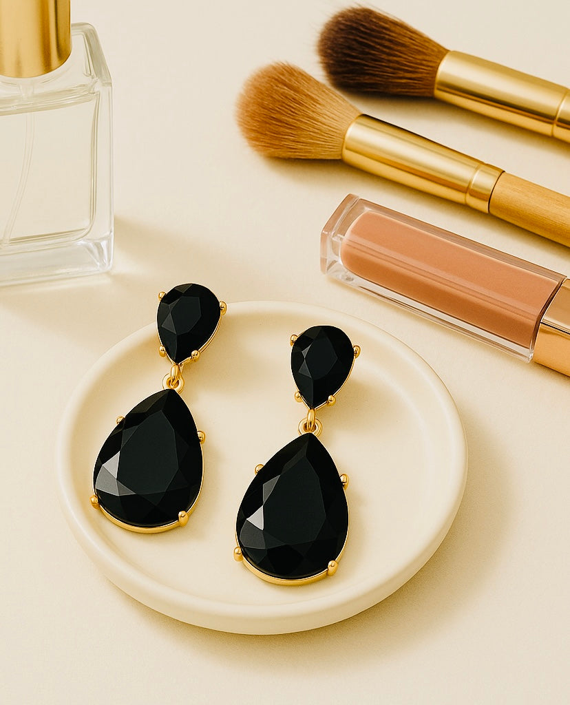 Black teardrop earrings on a white dish with makeup brushes and lip gloss in the background.