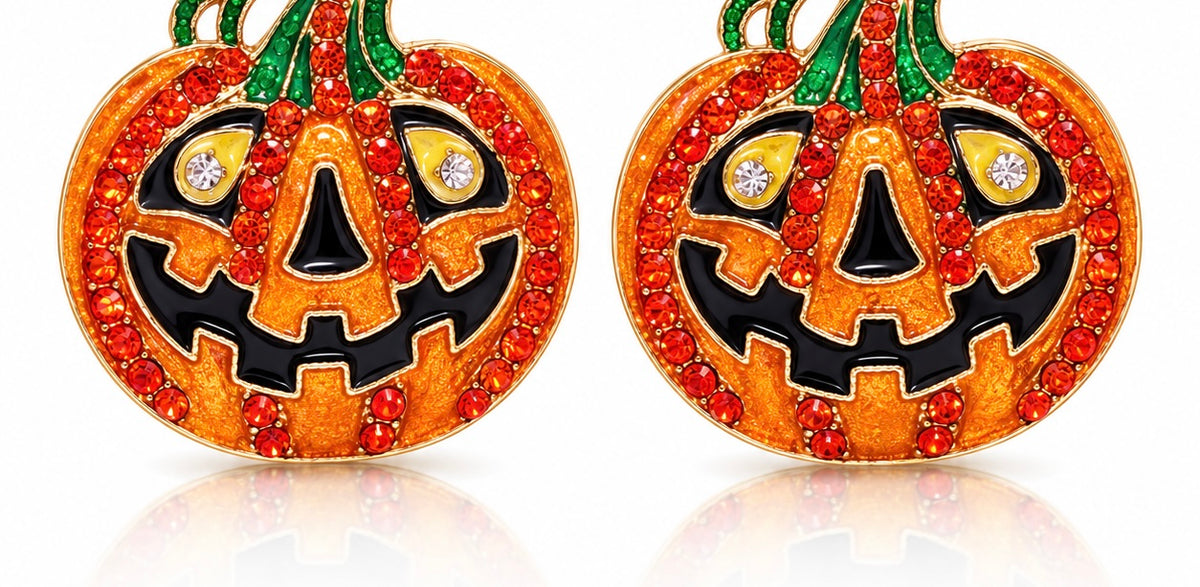 A pair of pumpkin-shaped earrings decorated with orange and black enamel, accented by red rhinestones, green stems, and clear crystal eyes, shown on a white background with a soft reflection.
