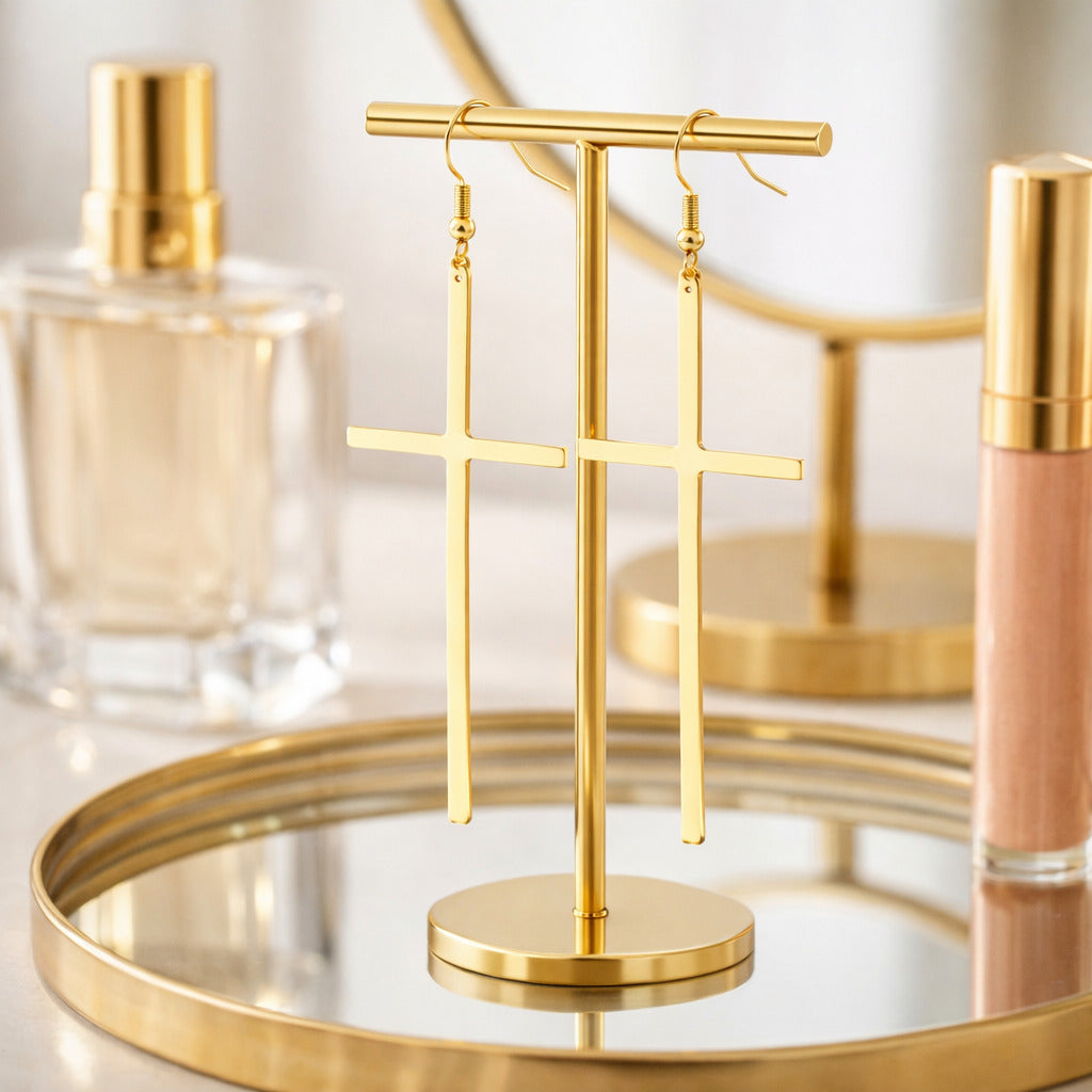 Gold earring stand with cross-shaped earrings on a reflective surface with cosmetic products in the background.