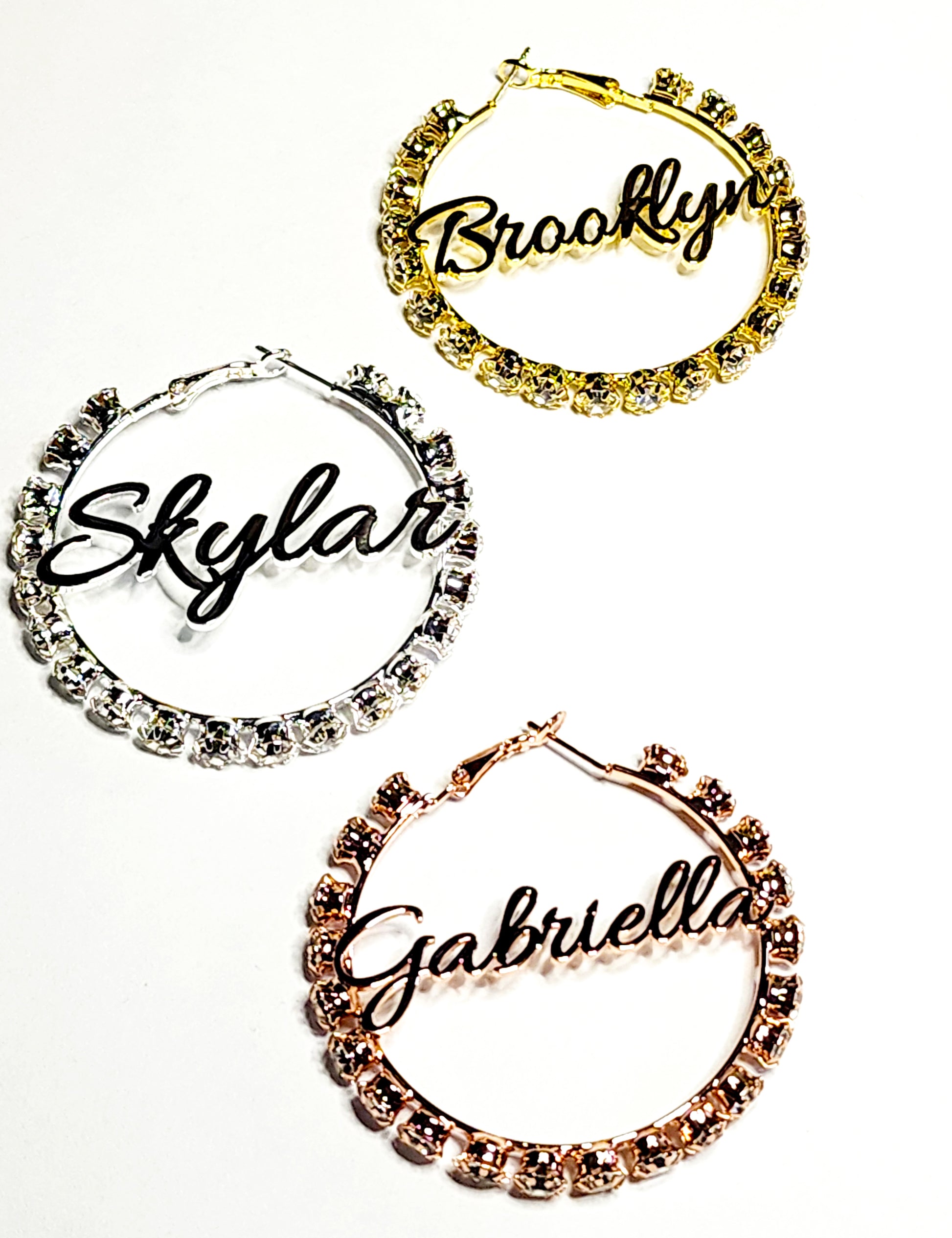 Three customized rhinestone hoop earrings in silver, gold, and rose gold colors, each with a different name written on them.