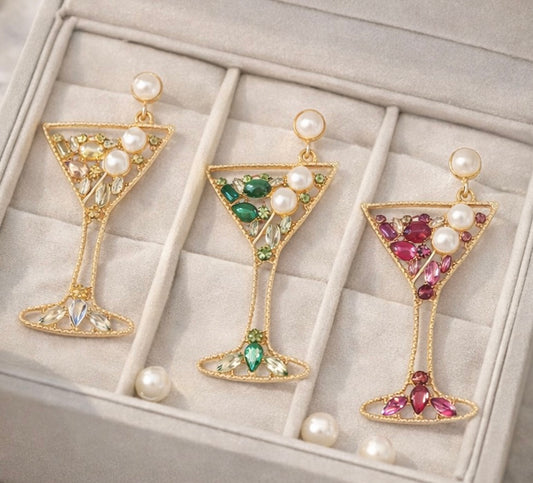 Three pairs of martini glass-shaped earrings with gemstones and pearls on a textured surface.