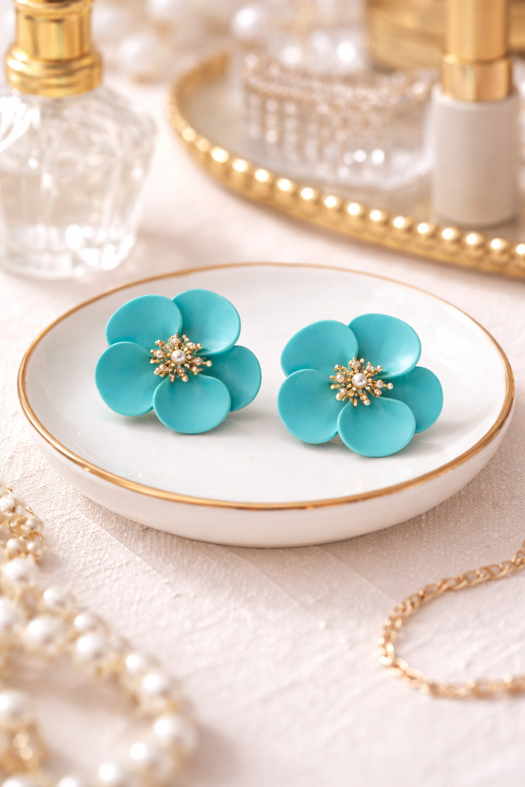 Turquoise flower earrings on a white dish with gold rim, pearl necklace, and mirror in the background.