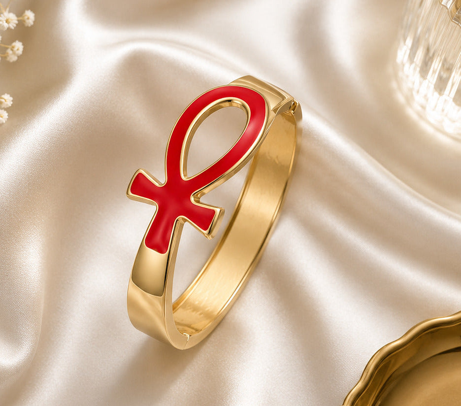A gold-plated cuff bracelet featuring a prominent red enamel Ankh symbol, styled on draped champagne satin with small white baby's breath flowers and a glass tumbler.
