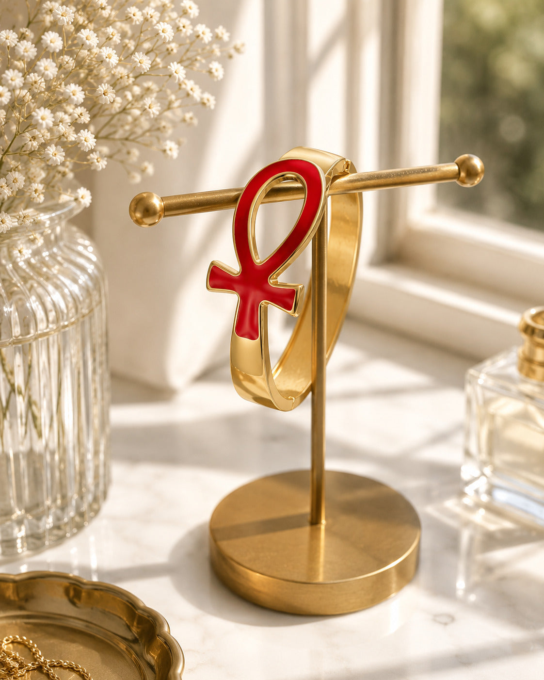 A gold ring stand holding a gold ring with a red ankh symbol, styled on a marble surface near a window with dried flowers and perfume bottles.