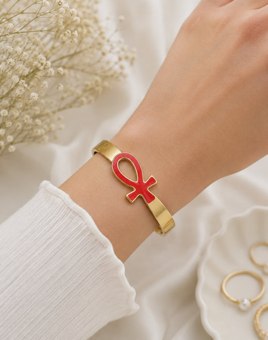 A gold-tone cuff bracelet with a red enamel Ankh symbol, worn on a person's wrist against a soft white fabric background with dried flowers.