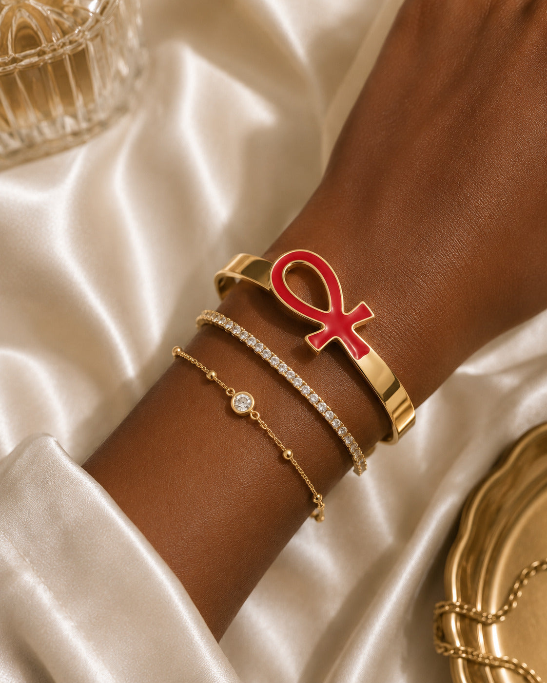 A close-up of a person's wrist wearing three gold bracelets: a wide cuff with a bold red ankh symbol, a slim bracelet with clear stones, and a delicate chain with a small round clear stone. The wrist rests on draped white satin fabric.