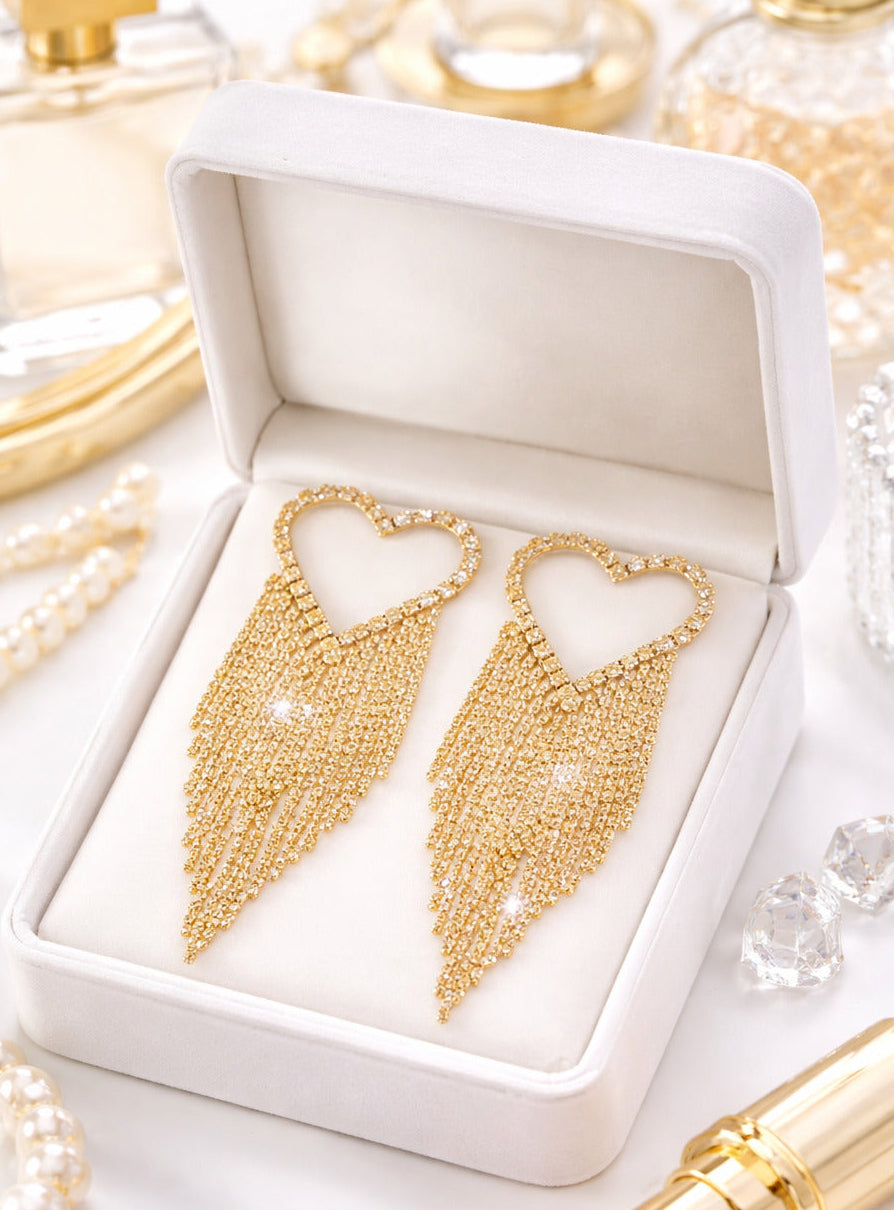 Gold heart-shaped earrings in a white box with perfume bottles and a lipstick in the background.