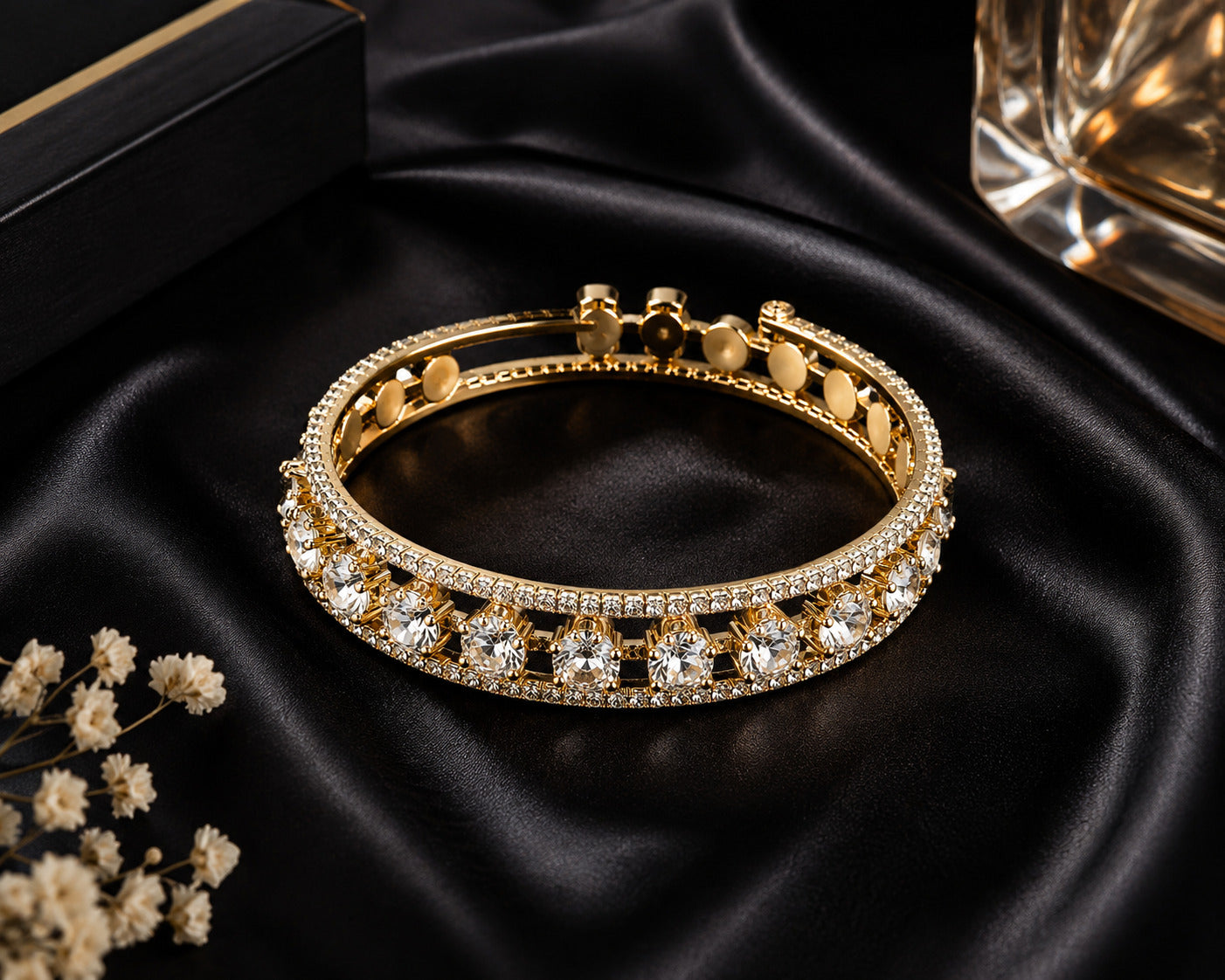 A gold-plated hinged bangle bracelet embellished with rows of clear crystals, displayed on a black satin fabric background with decorative dried flowers and a black box.