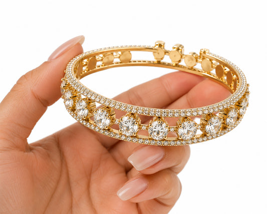 A gold bangle bracelet with pavé-set clear stones and large round center stones, held in a hand against a white background.