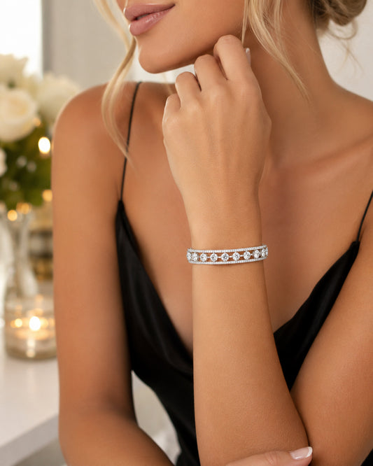 A close-up lifestyle shot of a woman wearing a slim silver-toned bracelet adorned with sparkling clear gemstones, set against a softly blurred indoor background with warm lighting.