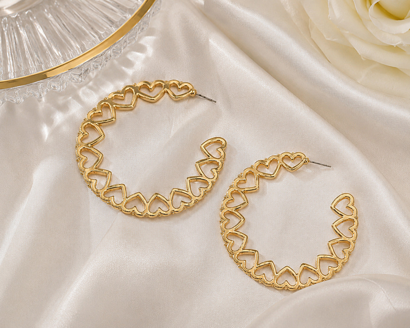 A pair of gold hoop earrings with an openwork heart-link design, displayed on draped white satin fabric next to a white rose and a glass dish.